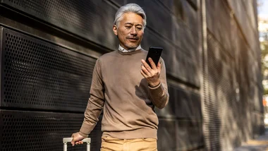 Man looking at a smartphone while pulling a suitcase behind him while he walks.